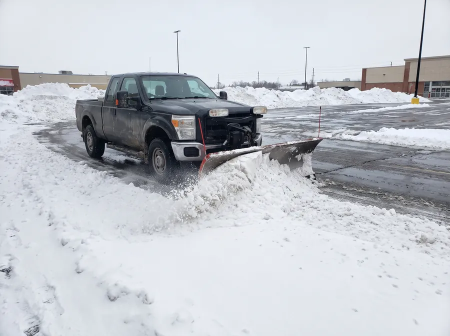Pickup truck with a front plow blade clearing snow across a commercial parking lot