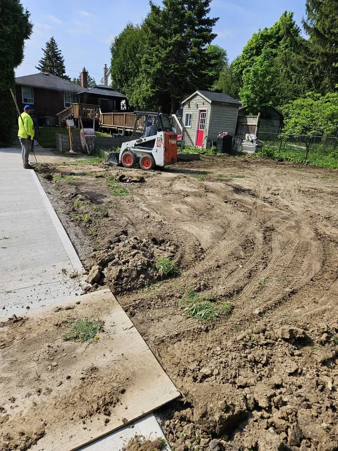Compact skid steer re-grading soil across a stripped yard beside a new concrete walkway