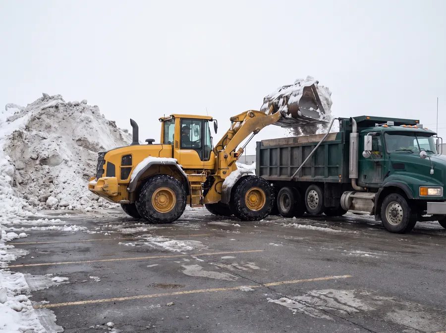 Wheel loader loading snow into a tandem dump truck beside a snow stockpile