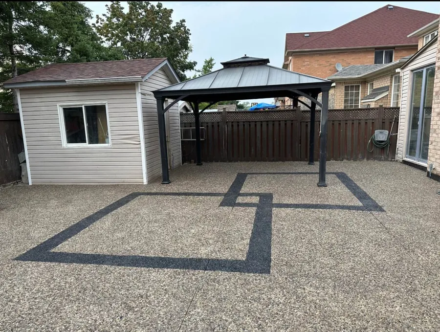 Exposed aggregate concrete patio with contrasting dark border bands surrounding a metal gazebo and garden shed.