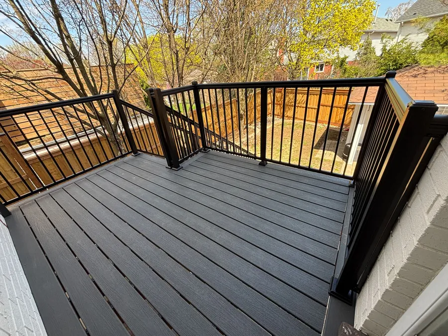Grey capped-composite deck boards with a black aluminum railing, sunlit, beside a new wood privacy fence.